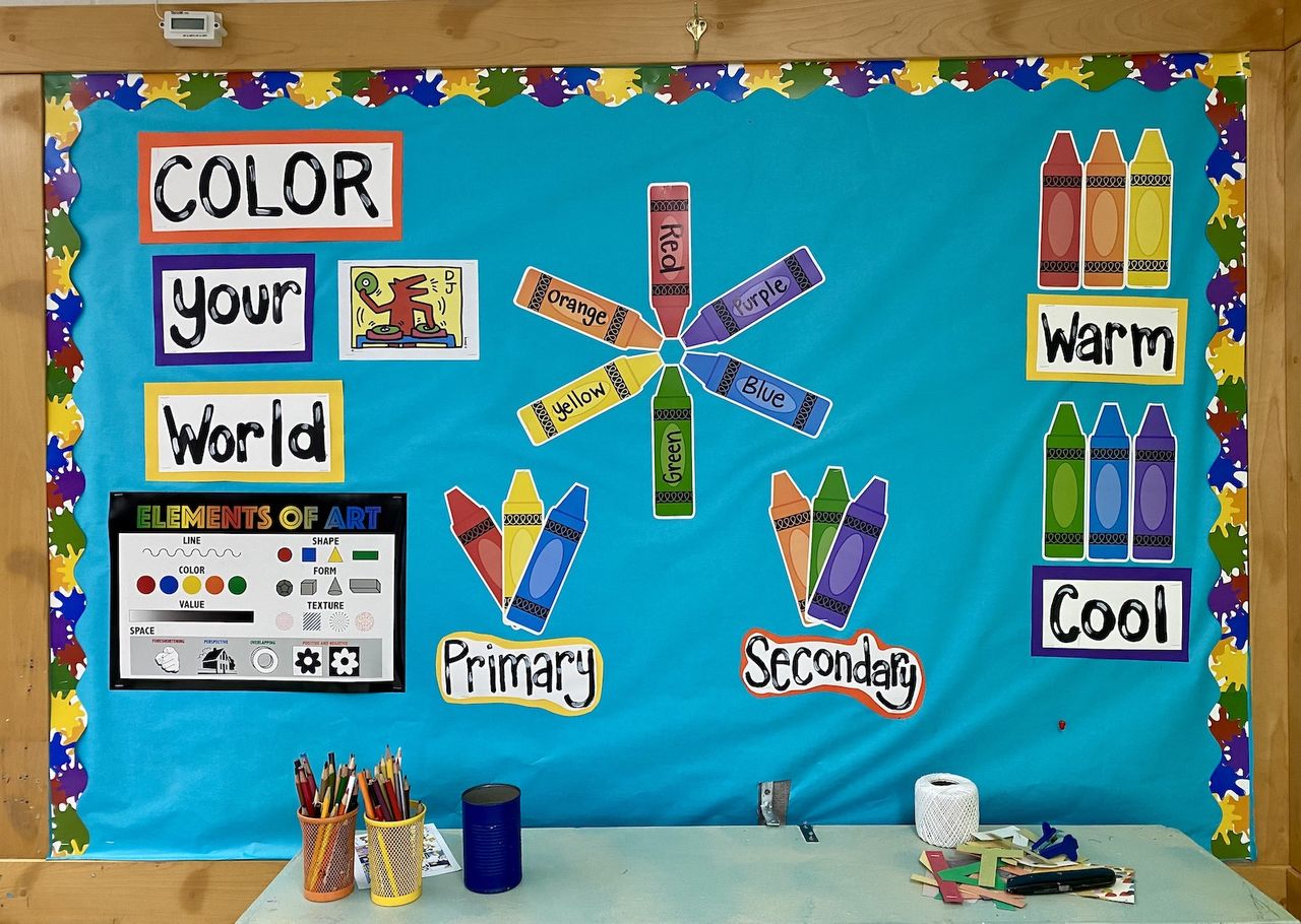 A classroom sign with a blue background titled Color Your World. Large crayons in groups are being used to show primary versus secondary colors, and warn versus cool colors.