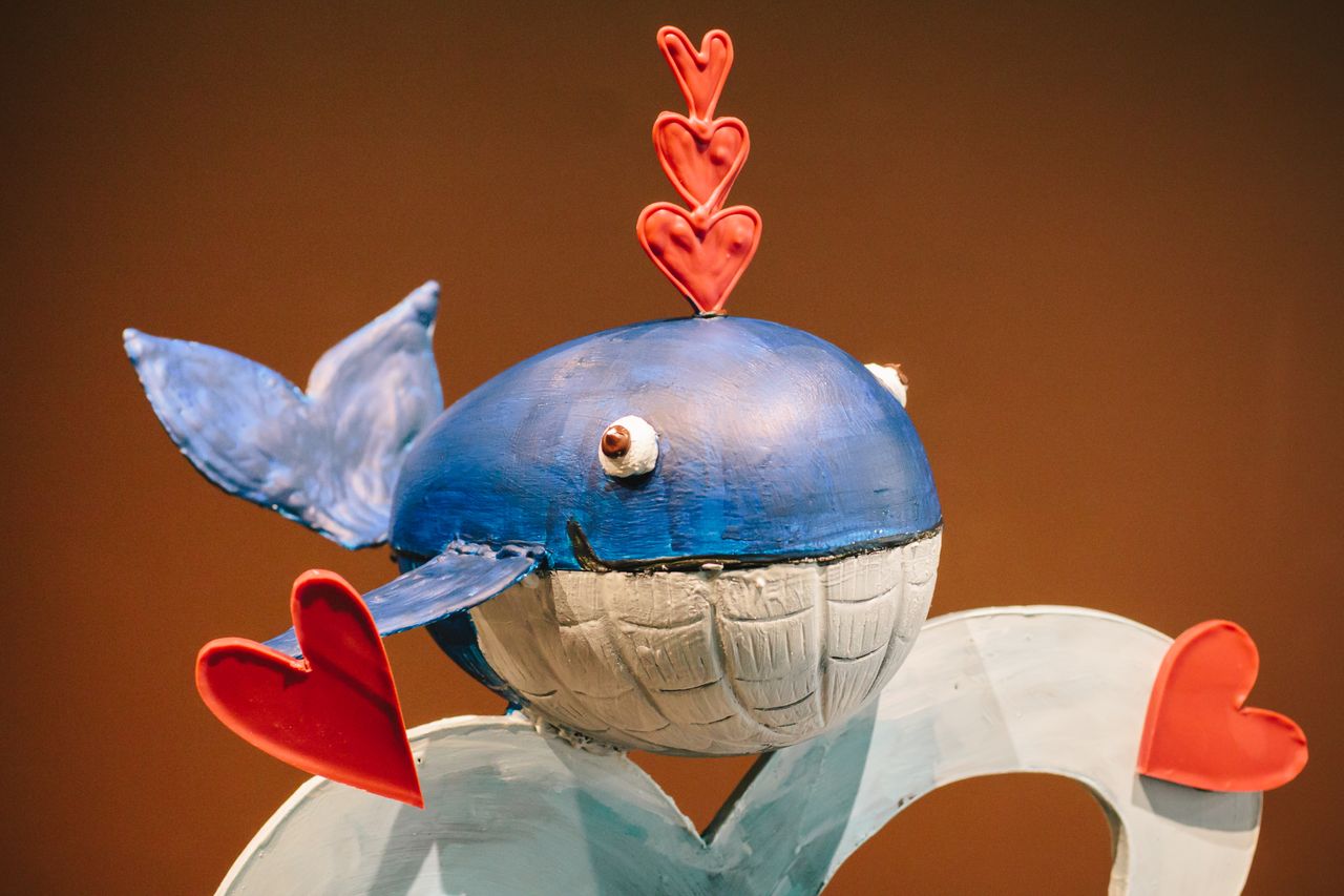 A close-up of a smiling chocolate whale, painted with blue colored cocoa butter, with 3 red hearts coming out of its spout.