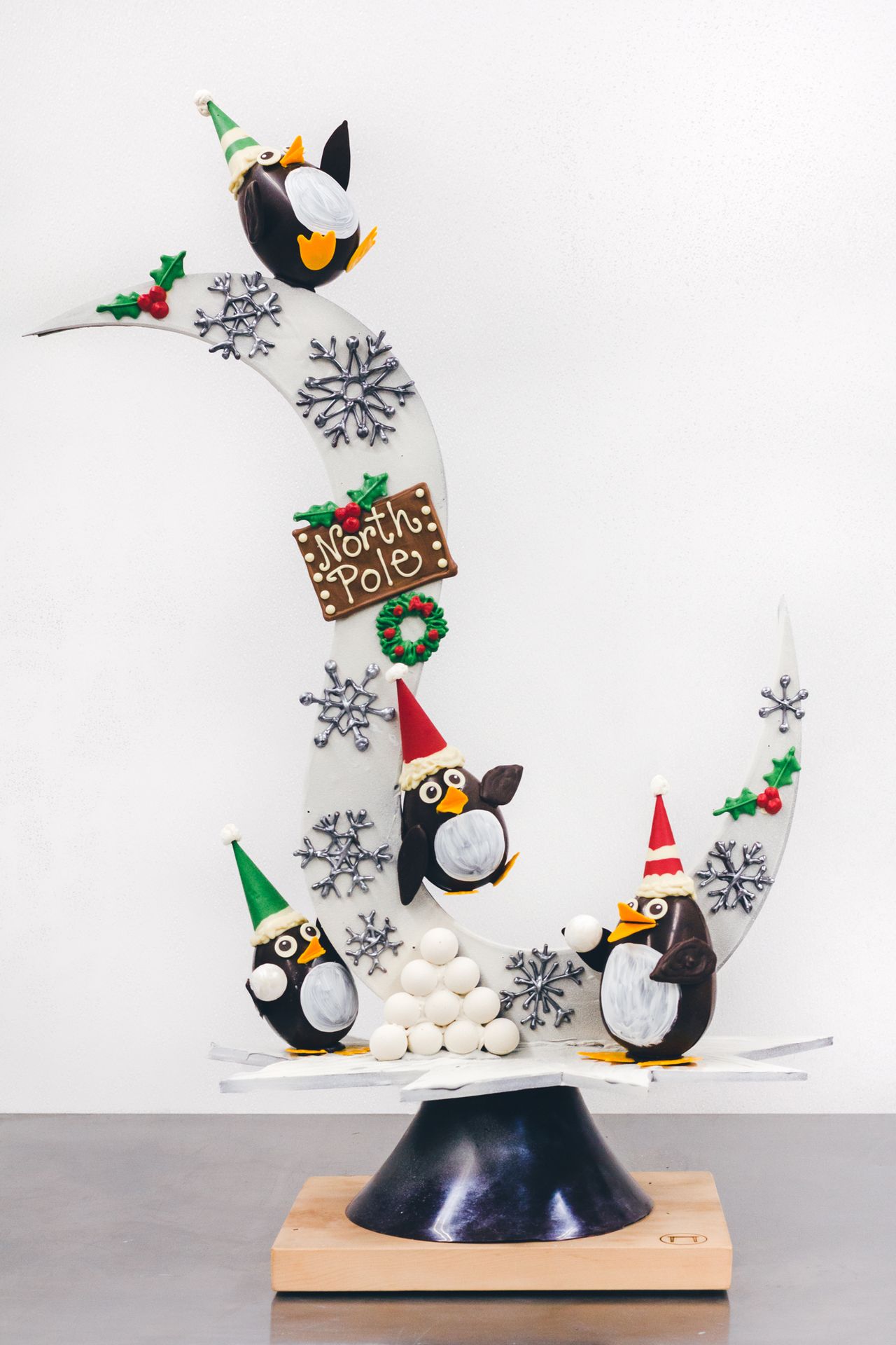 A large chocolate Christmas sculpture with cute penguins wearing elves hats having a snowball fight.