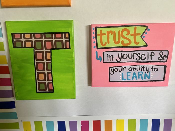 Two painted canvases, on the left a large letter T on a green background. On the right the phrase 'Trust in yourself and your ability to learn'